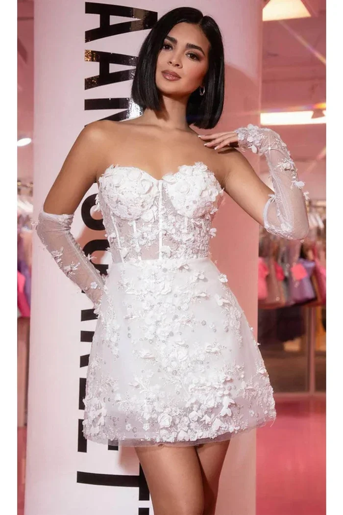 A woman wearing a white, strapless, floral 3D dress with long lace gloves posing indoors with a pink background and a mannequin display behind her.