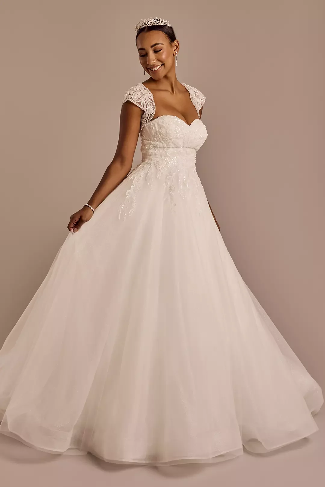 Woman in a white wedding gown with lace details, wearing a tiara and earrings, smiling.