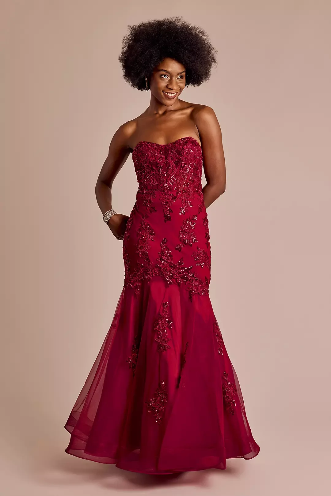 A woman wearing a strapless, floor-length red gown with floral lace embroidery and beadwork, smiling and standing against a neutral background.