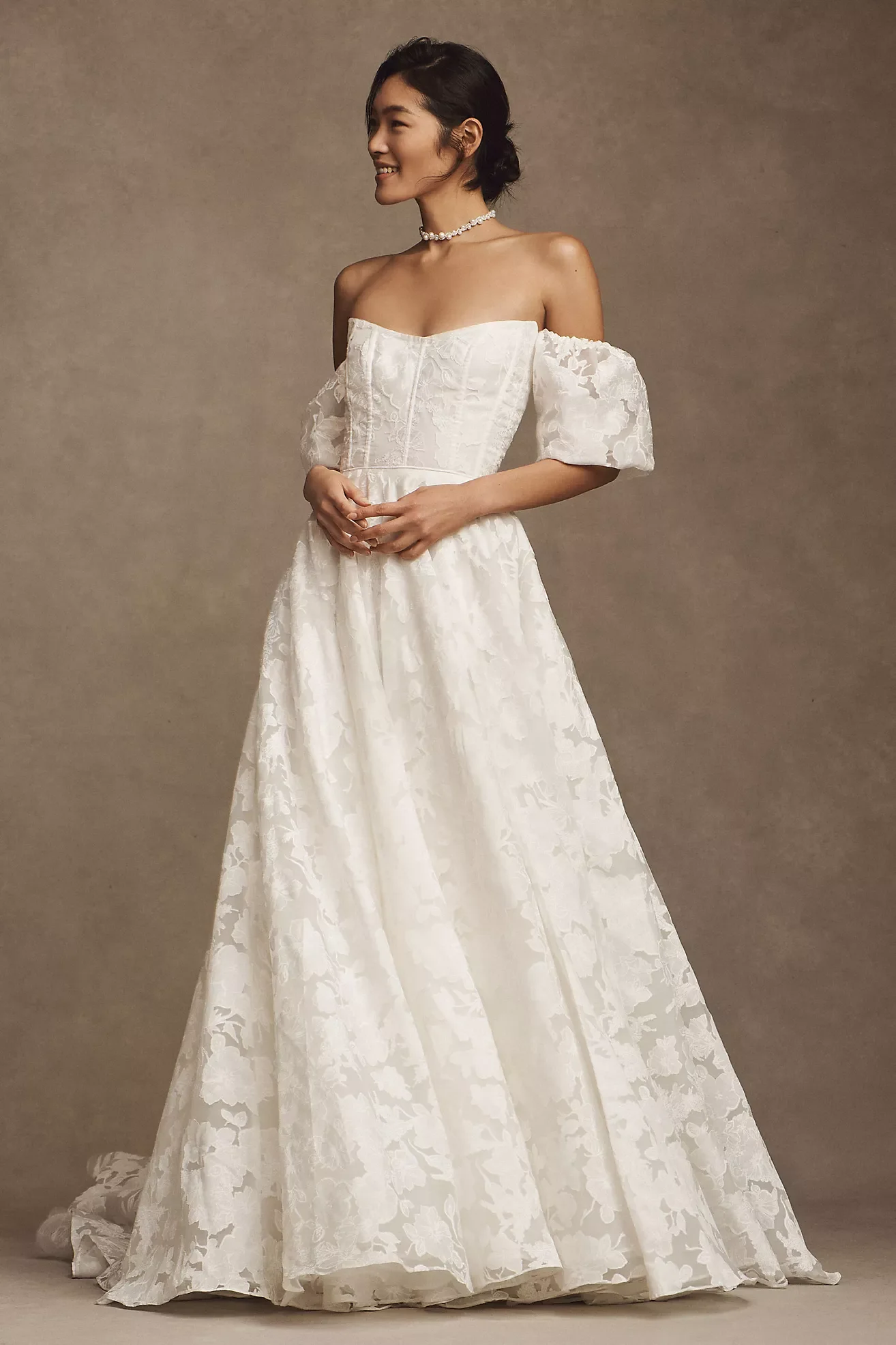 A woman wearing a strapless white wedding gown with a floral pattern, standing in front of a neutral brown background, smiling and holding her hands together at her waist, accessorized with a pearl necklace.