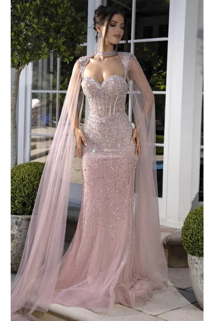 A woman in a glittery, pink evening gown with sheer, dramatic cape sleeves stands outdoors near potted plants and glass doors.