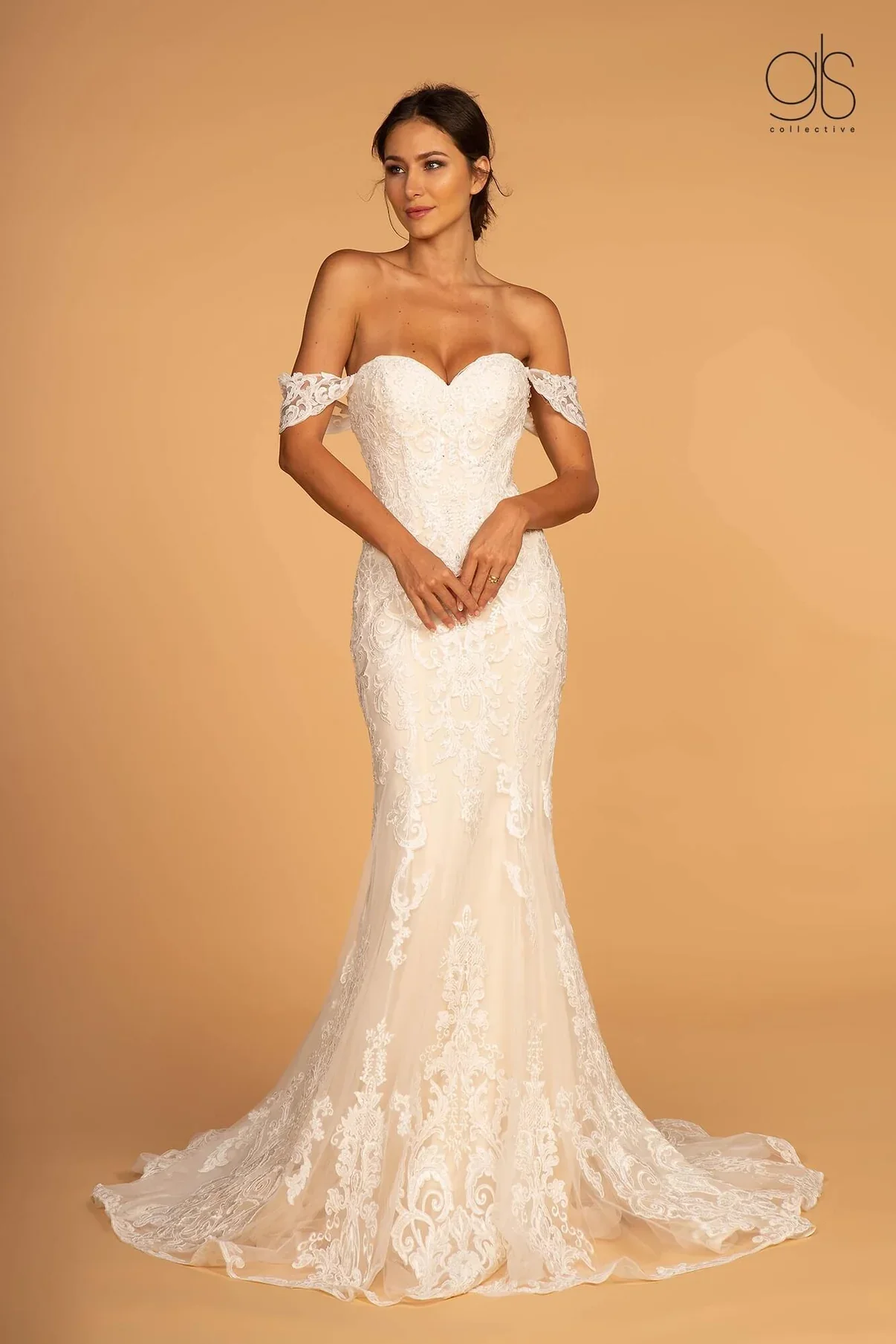 A woman wearing an off-the-shoulder white lace wedding dress with a fitted bodice and a mermaid silhouette, standing against a beige background.