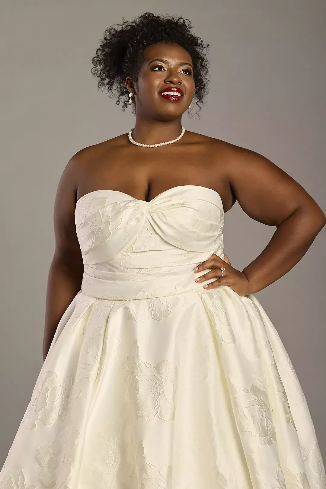 A woman dressed in a strapless cream-colored gown with floral patterns, wearing pearl jewelry, and standing with one hand on her hip, smiling against a neutral background.