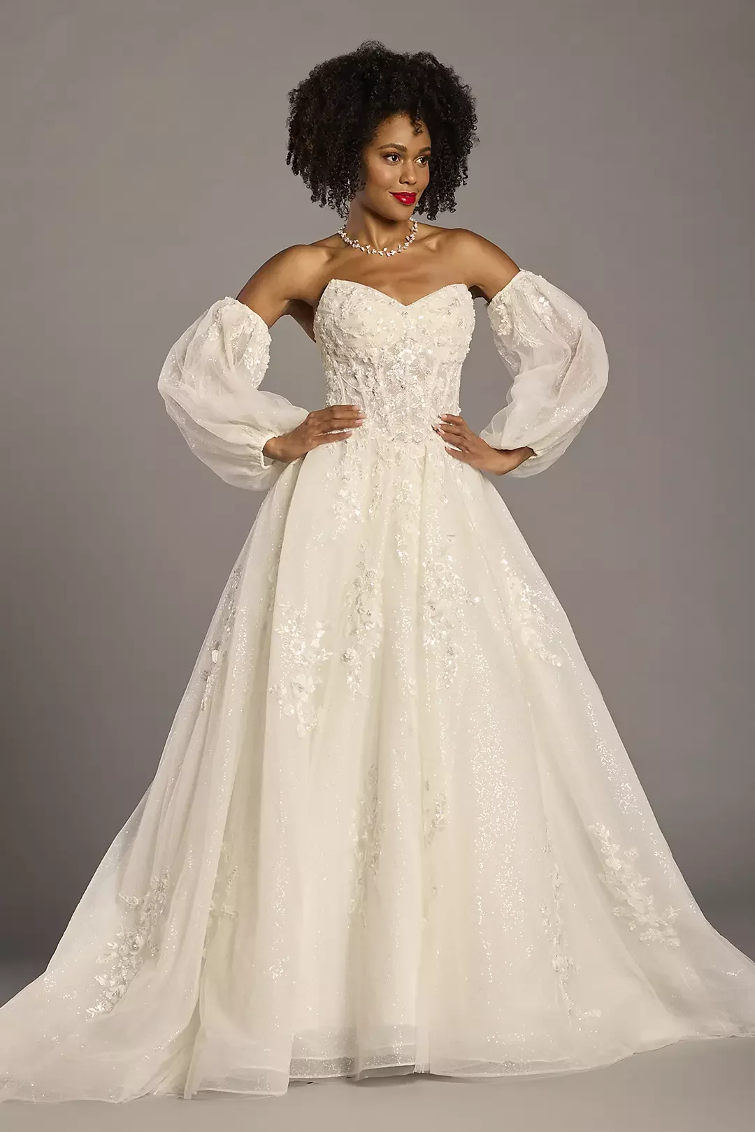 A woman modeling an elegant off-shoulder wedding dress with puffed long sheer sleeves, intricate embroidery, and a full-length flowing skirt, standing against a gray background.