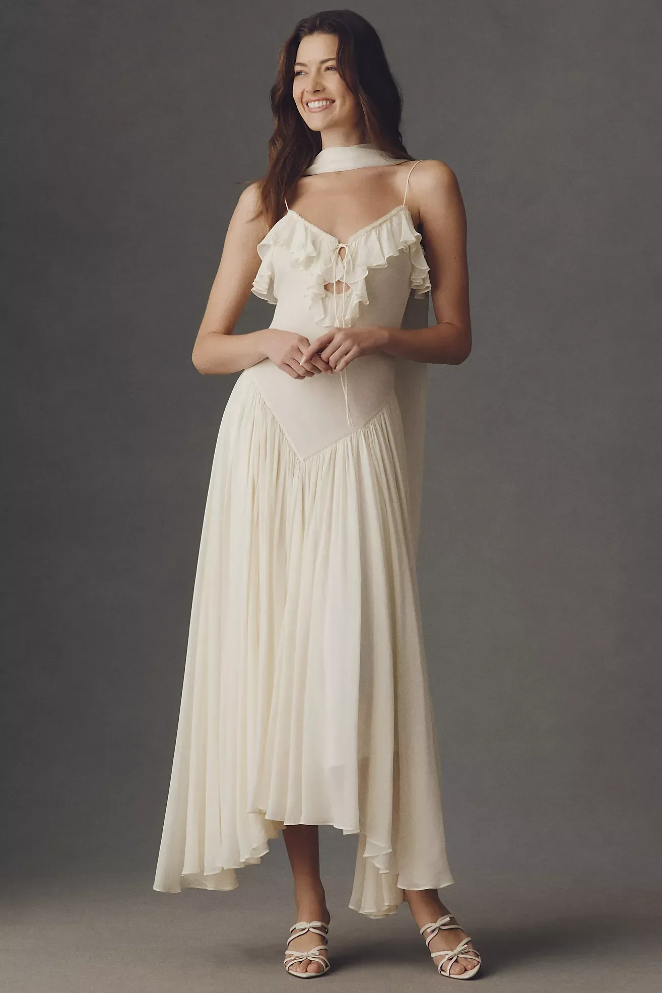 Woman in a cream-colored, flowy gown with ruffled top, standing against a gray background, smiling, wearing high heels with bows.