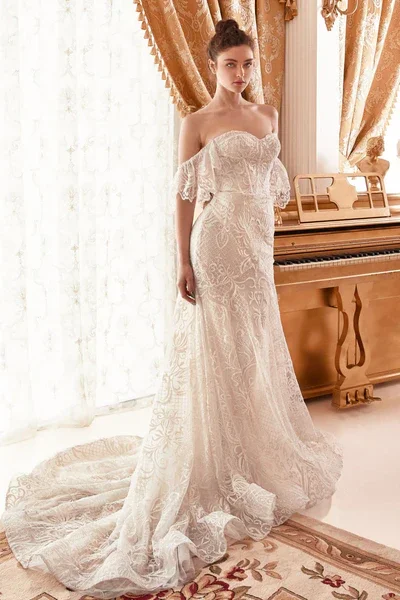 A woman in a strapless, off-the-shoulder, white lace wedding dress standing indoors next to a wooden piano, with gold curtains and sheer white curtains in the background.