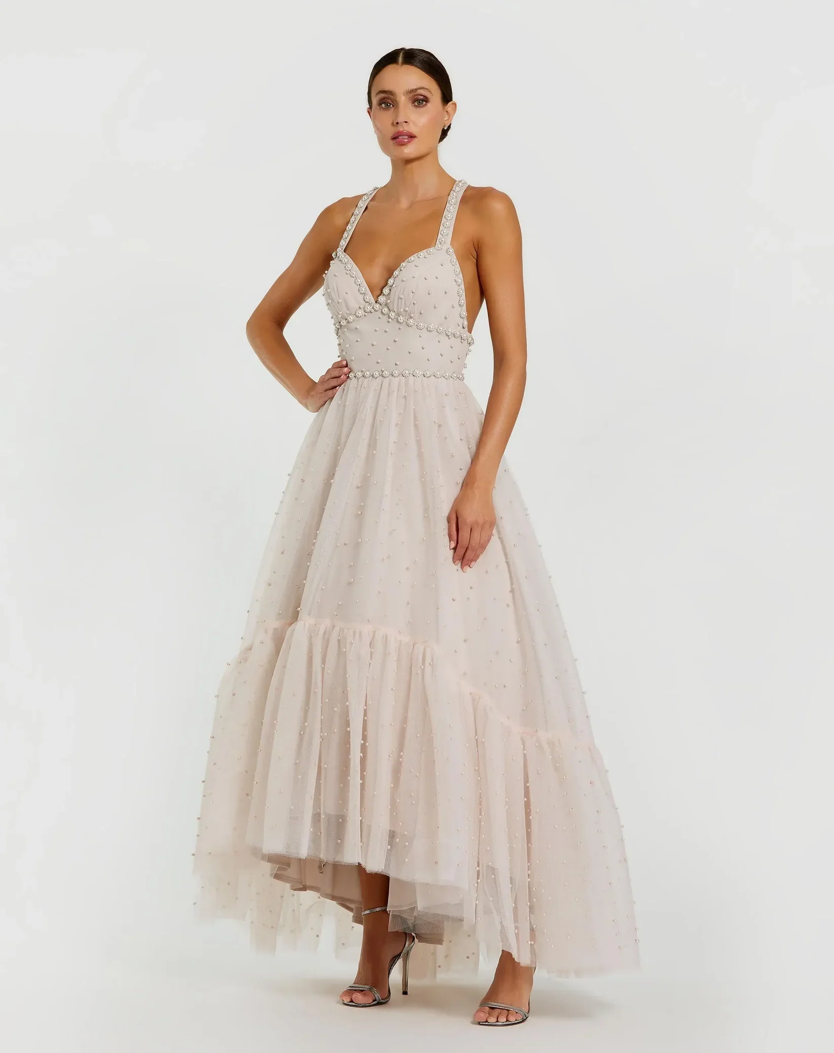 A woman in a light pink, sleeveless, floor-length dress with pearl embellishments, standing with one hand on her hip against a plain white background.