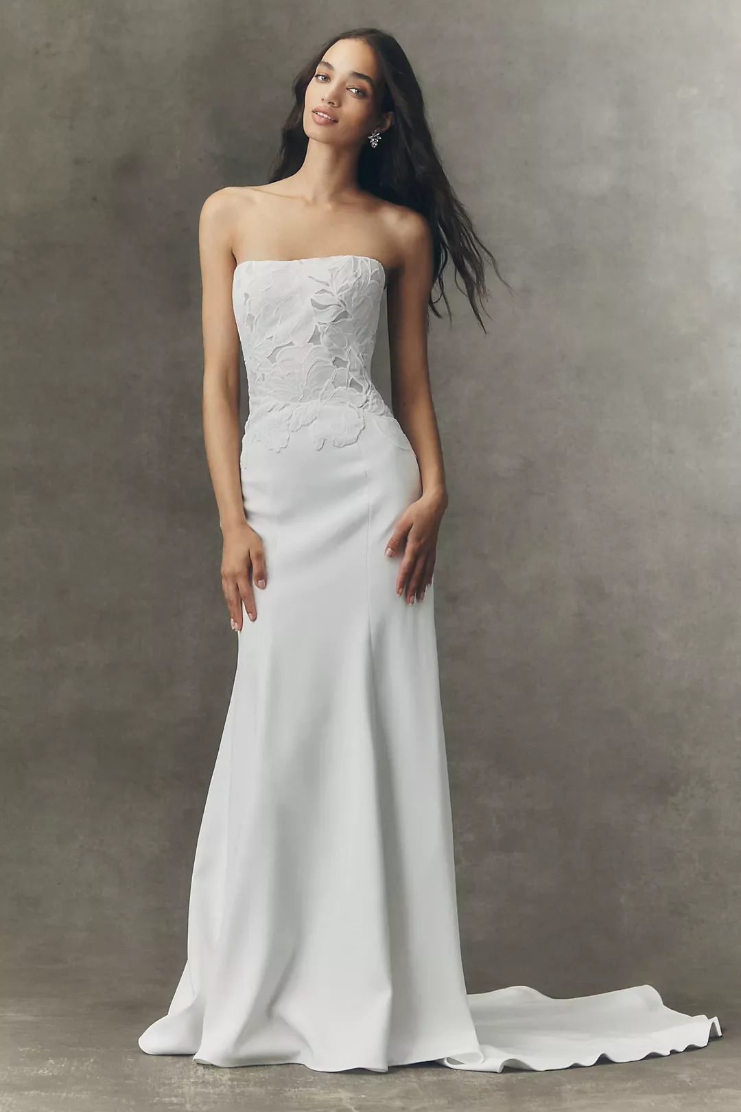 A woman wearing a strapless white wedding gown with a fitted bodice and floor-length skirt, standing against a plain background.