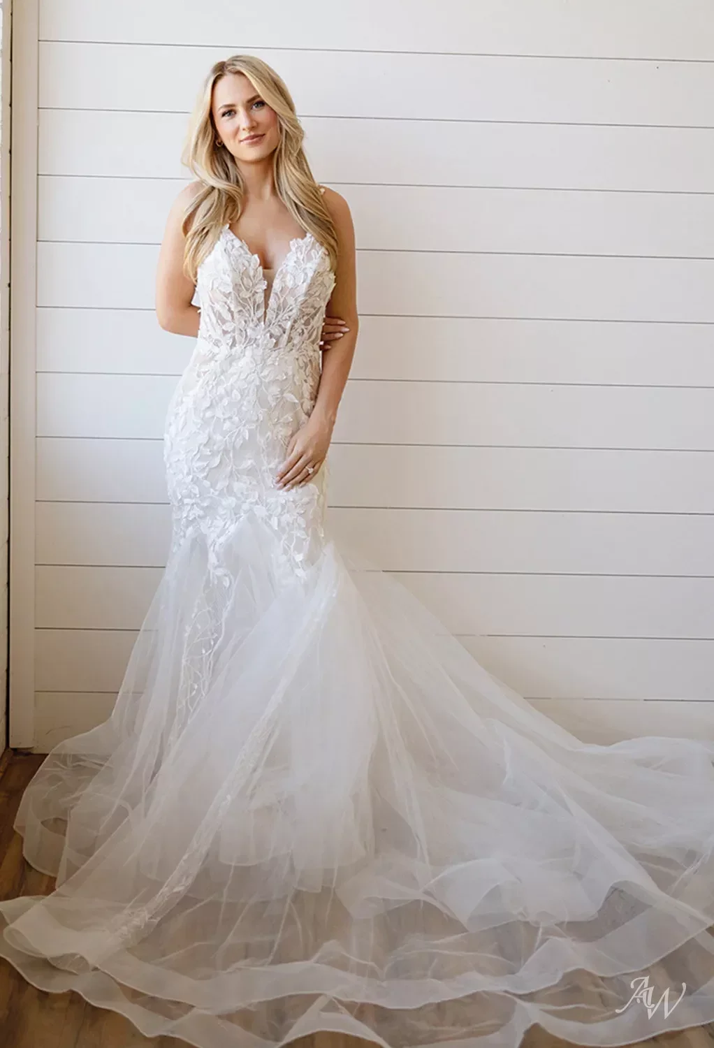 A woman in a white, lace wedding dress with a long tulle train stands against a white wooden wall.