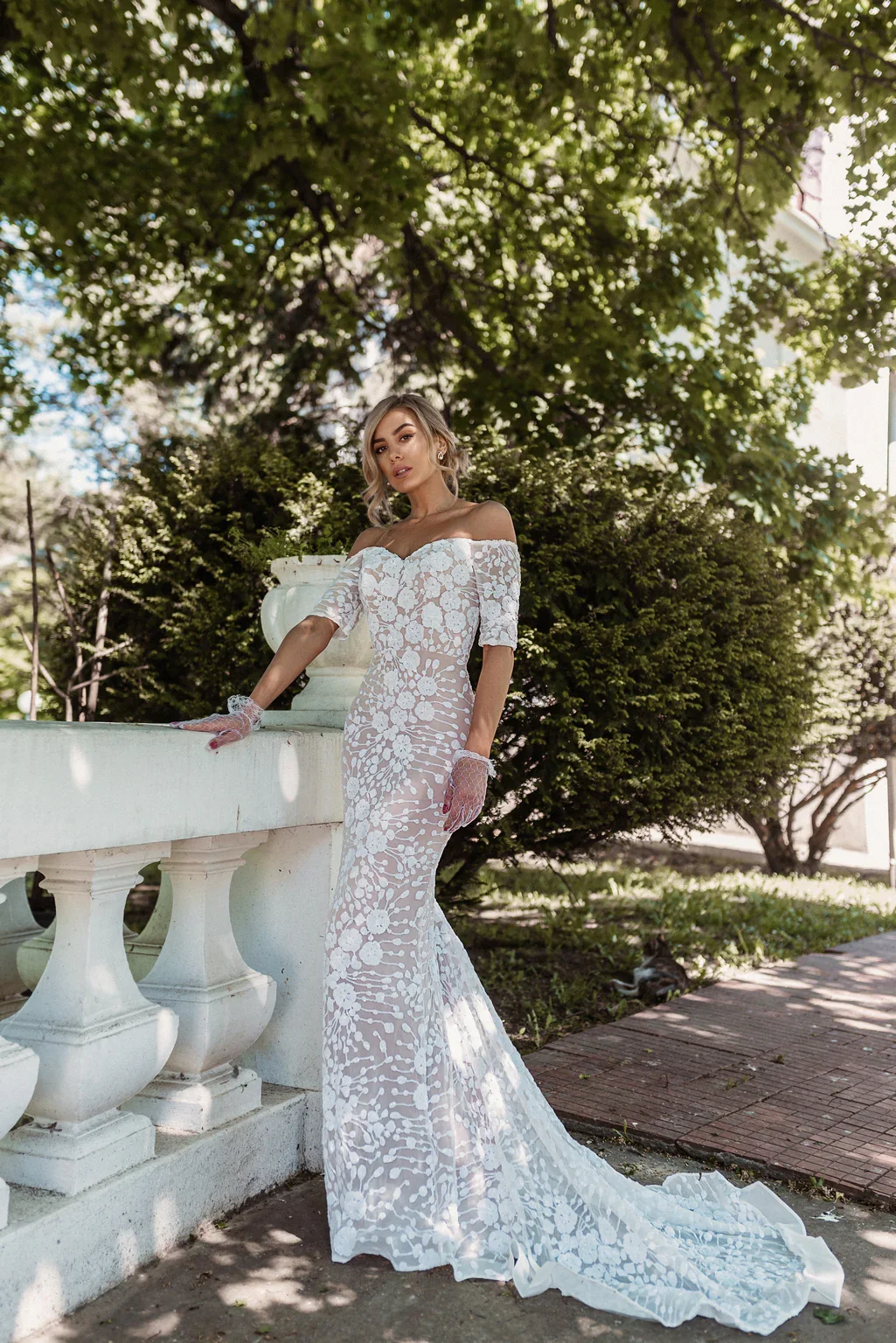 A woman in a white lace off-the-shoulder wedding dress with a train, standing outdoors near a white stone railing, with green trees and bushes in the background.