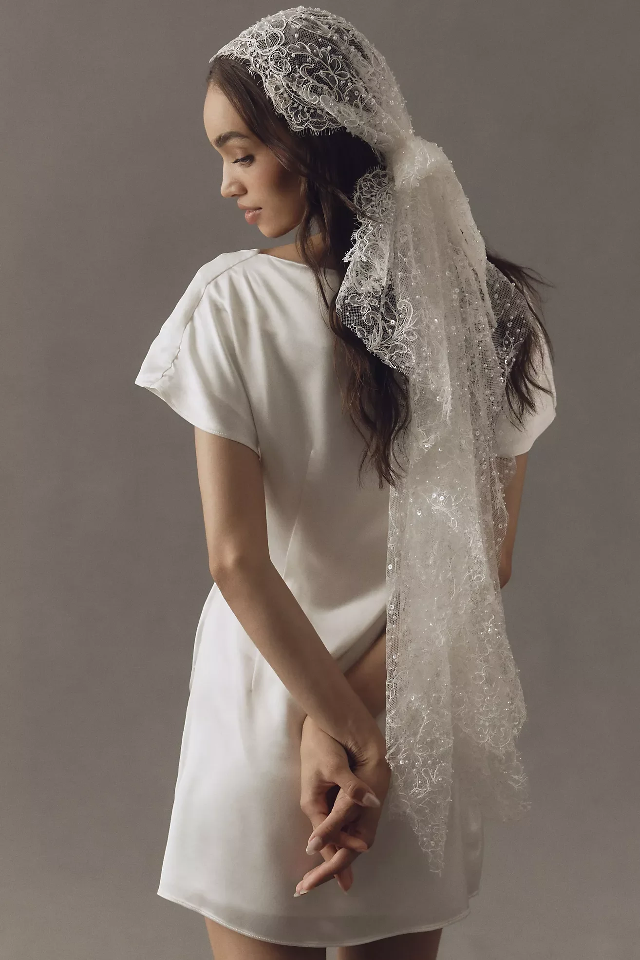 A woman with long dark hair wearing a white dress and a lace headscarf stands against a gray background, with her head slightly bowed and hands clasped.