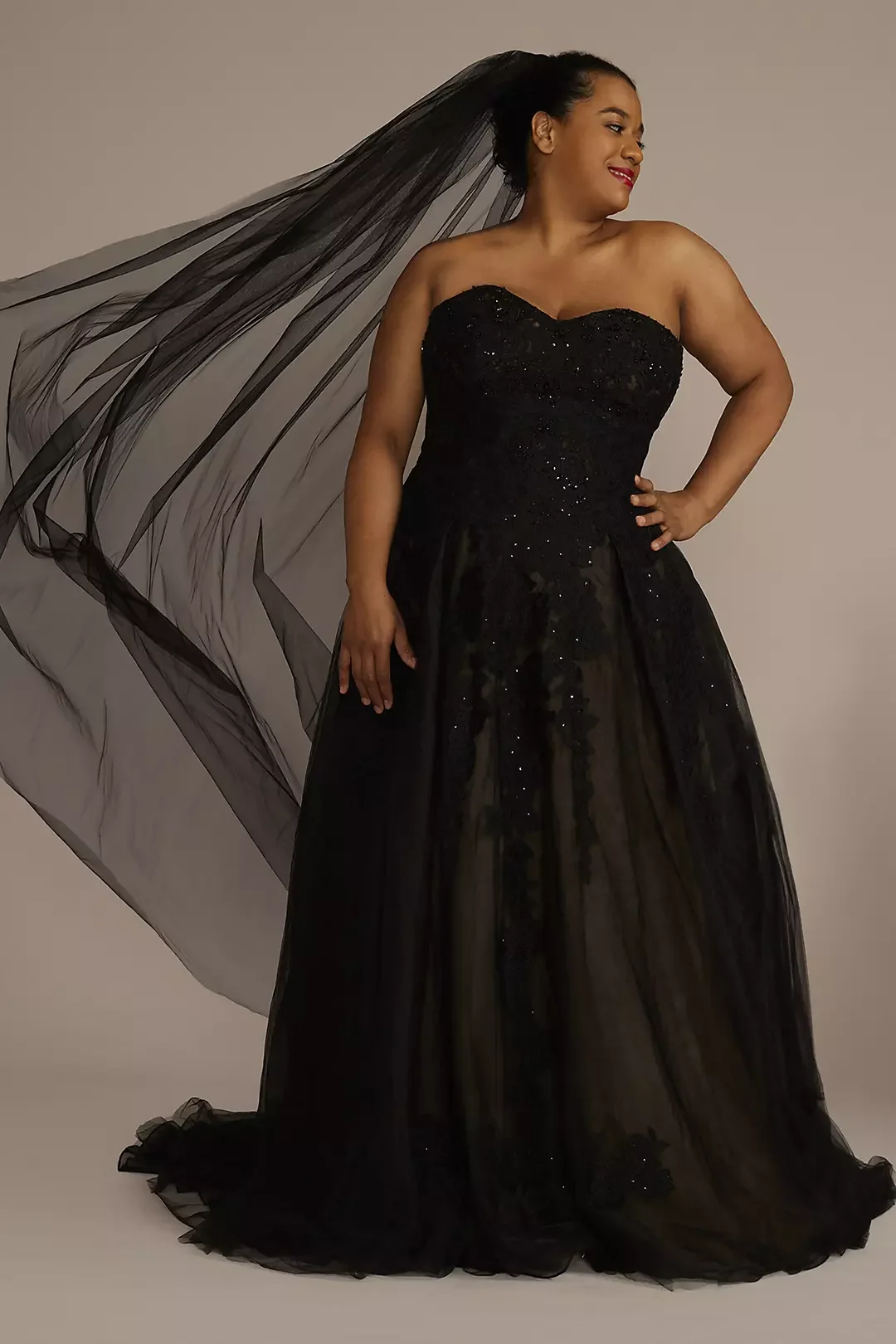 A woman in a black strapless, sequin-embellished gown with a sheer tulle overlay, standing against a neutral background, smiling and looking to the side.