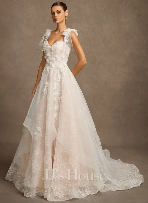 A woman wearing a white, sleeveless lace wedding gown with floral appliqués and a flowing train, standing against a neutral background.