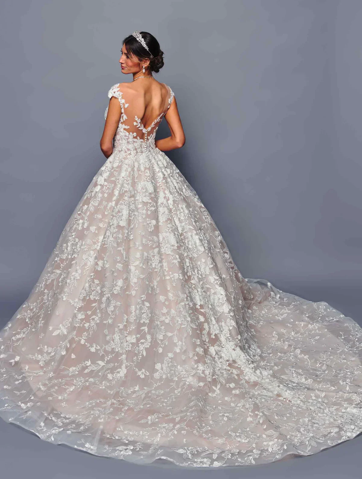 A woman wearing a white wedding gown with floral lace details and a sheer back, standing against a plain gray background.