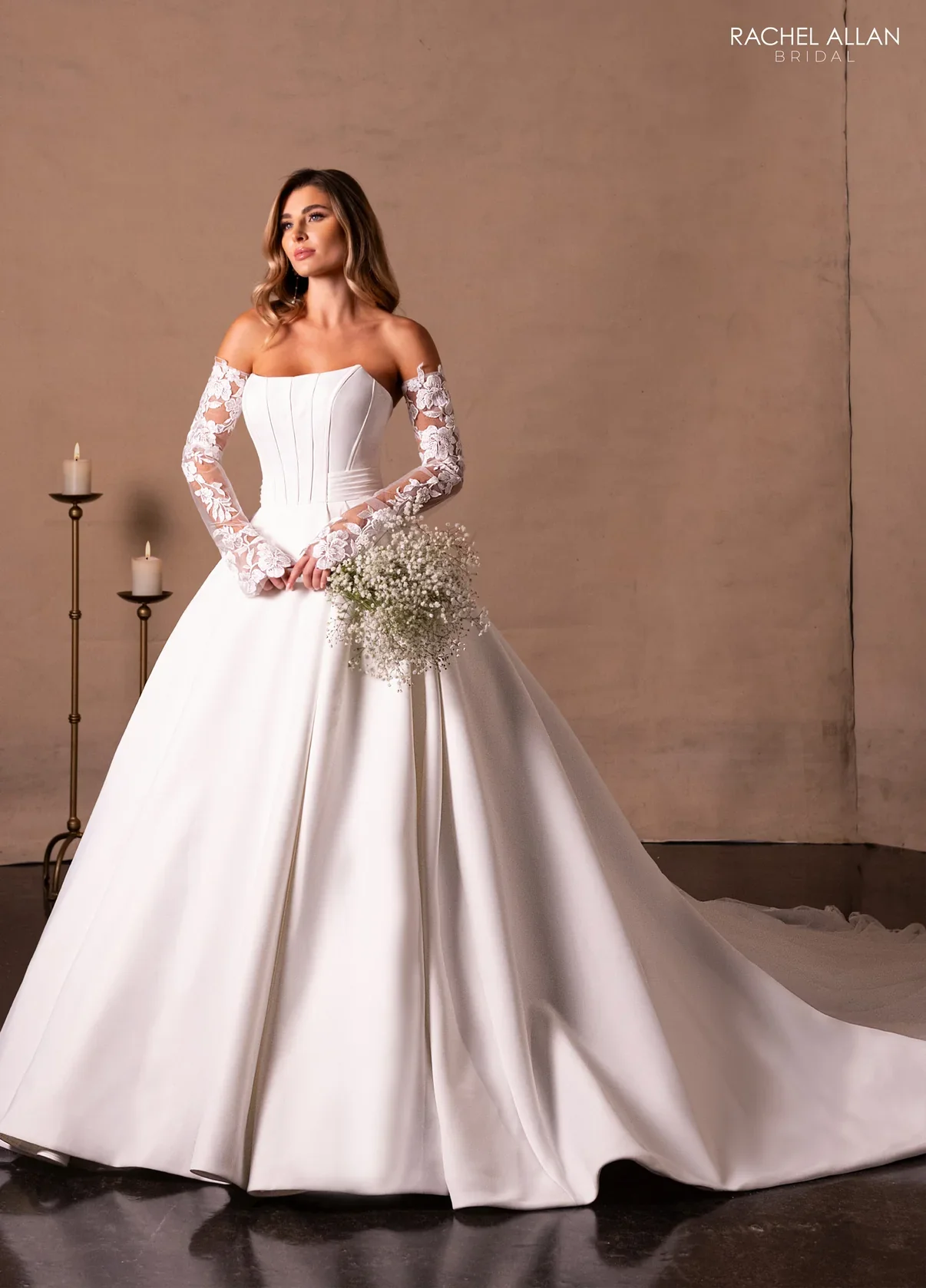 Bridal model in a white wedding gown with lace off-shoulder sleeves, holding a bouquet of baby's breath flowers, standing indoors against a plain backdrop with candles.