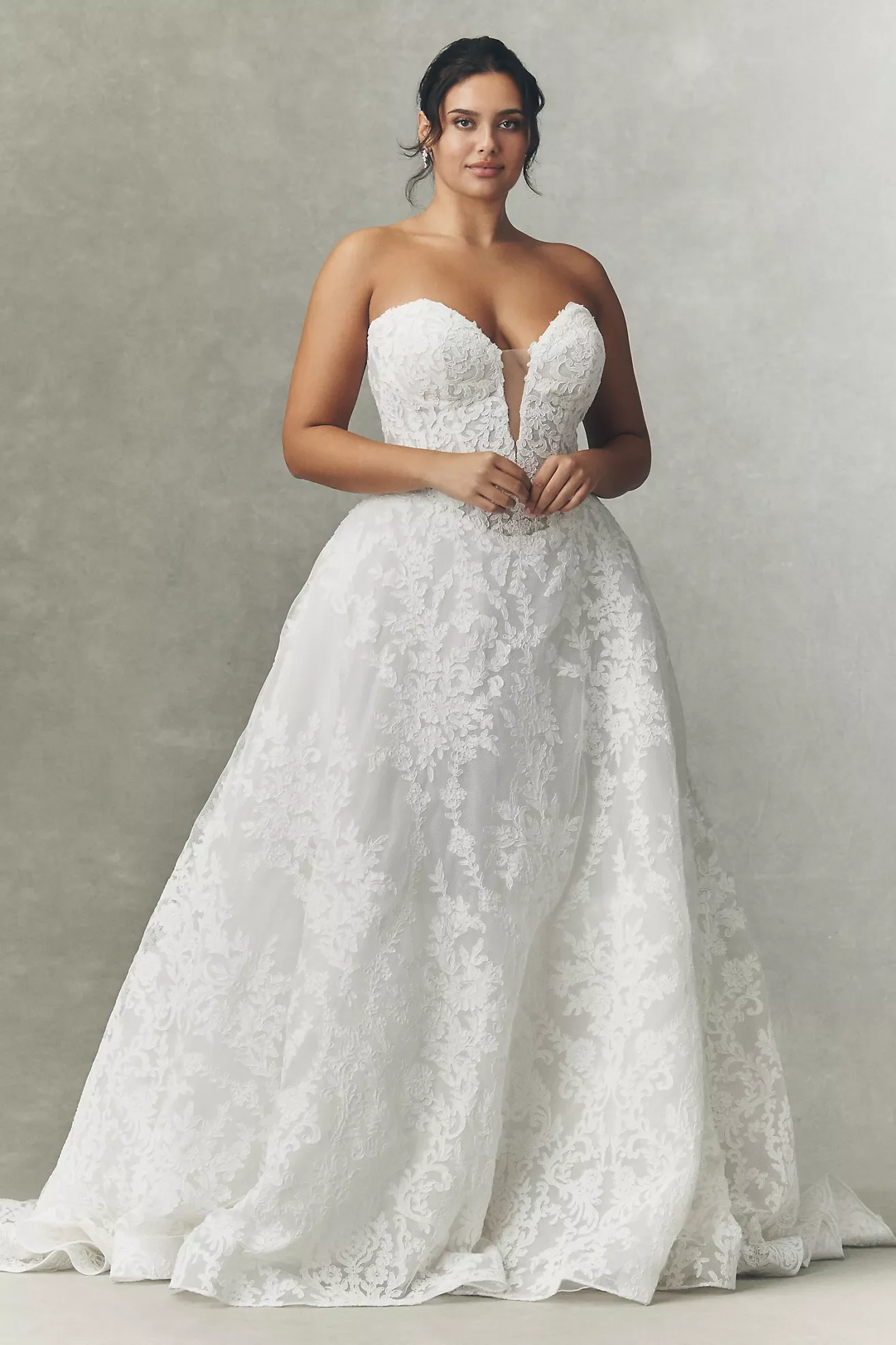 A woman in a strapless white wedding dress with lace appliqué details, standing against a plain light-colored background, with her hands gently clasped at her waist.