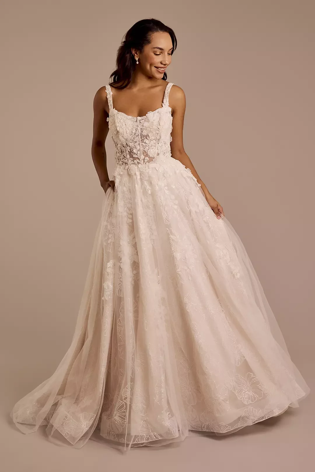 A woman wearing a white lace wedding dress with floral embroidery, a fitted bodice, and a full skirt, standing against a plain beige background.