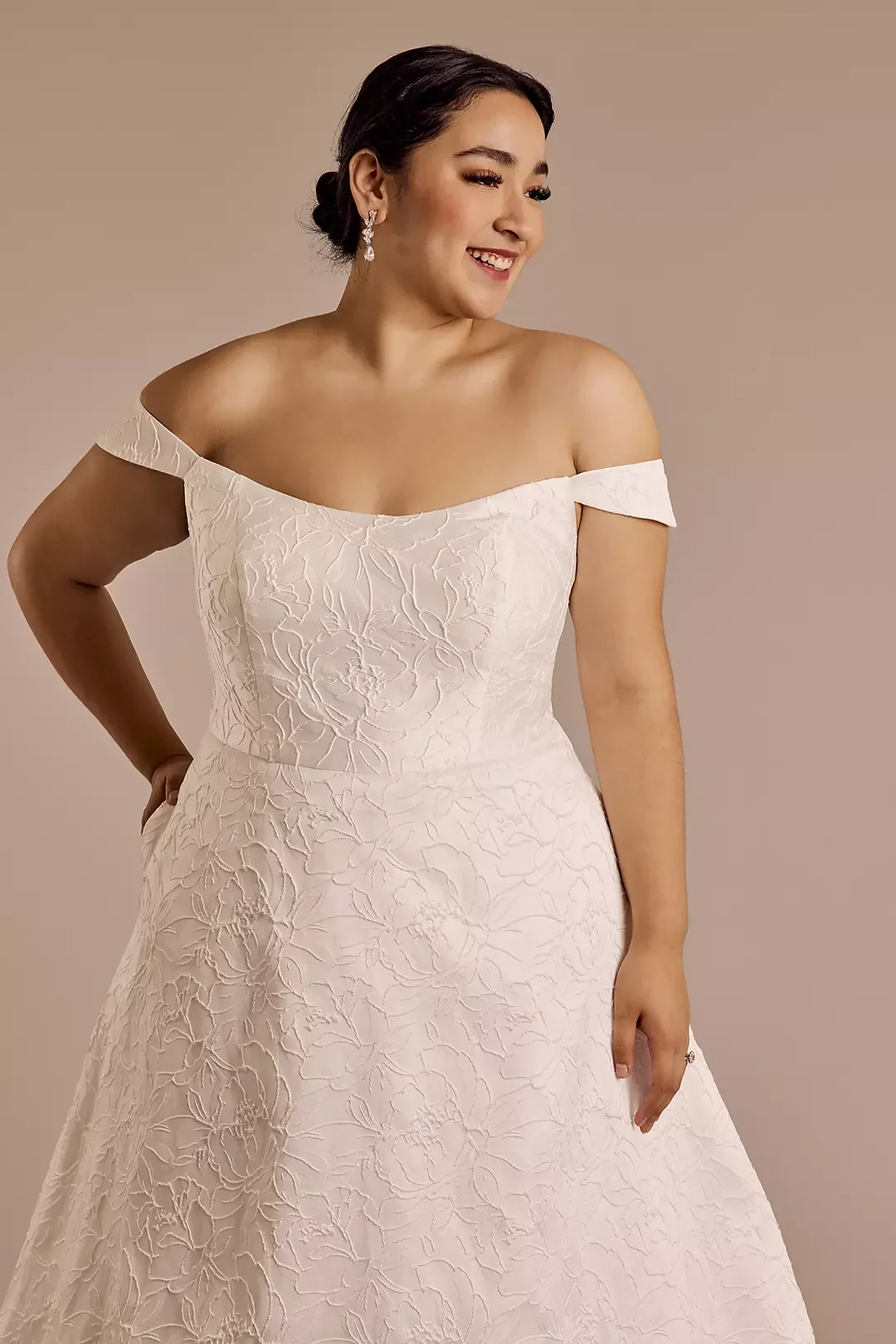 A woman smiling and looking to her right, wearing an off-shoulder white wedding dress with floral embroidery, and pearl earrings.