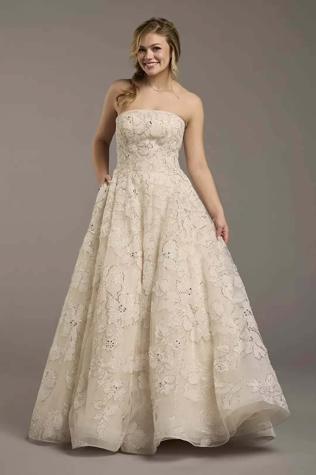 A young woman wearing an elegant strapless wedding dress with lace floral embroidery, standing against a plain grey background, smiling at the camera.