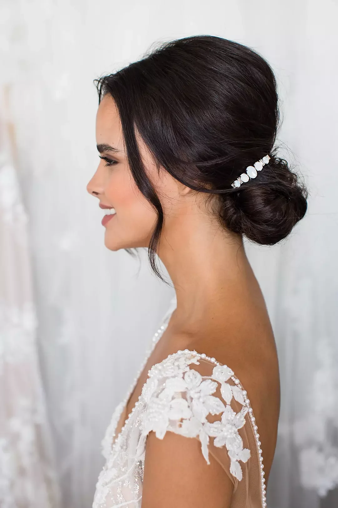Side profile of a woman with dark hair styled in an elegant updo with a decorative hairpiece, wearing a white lace dress with floral embroidery on the shoulder.
