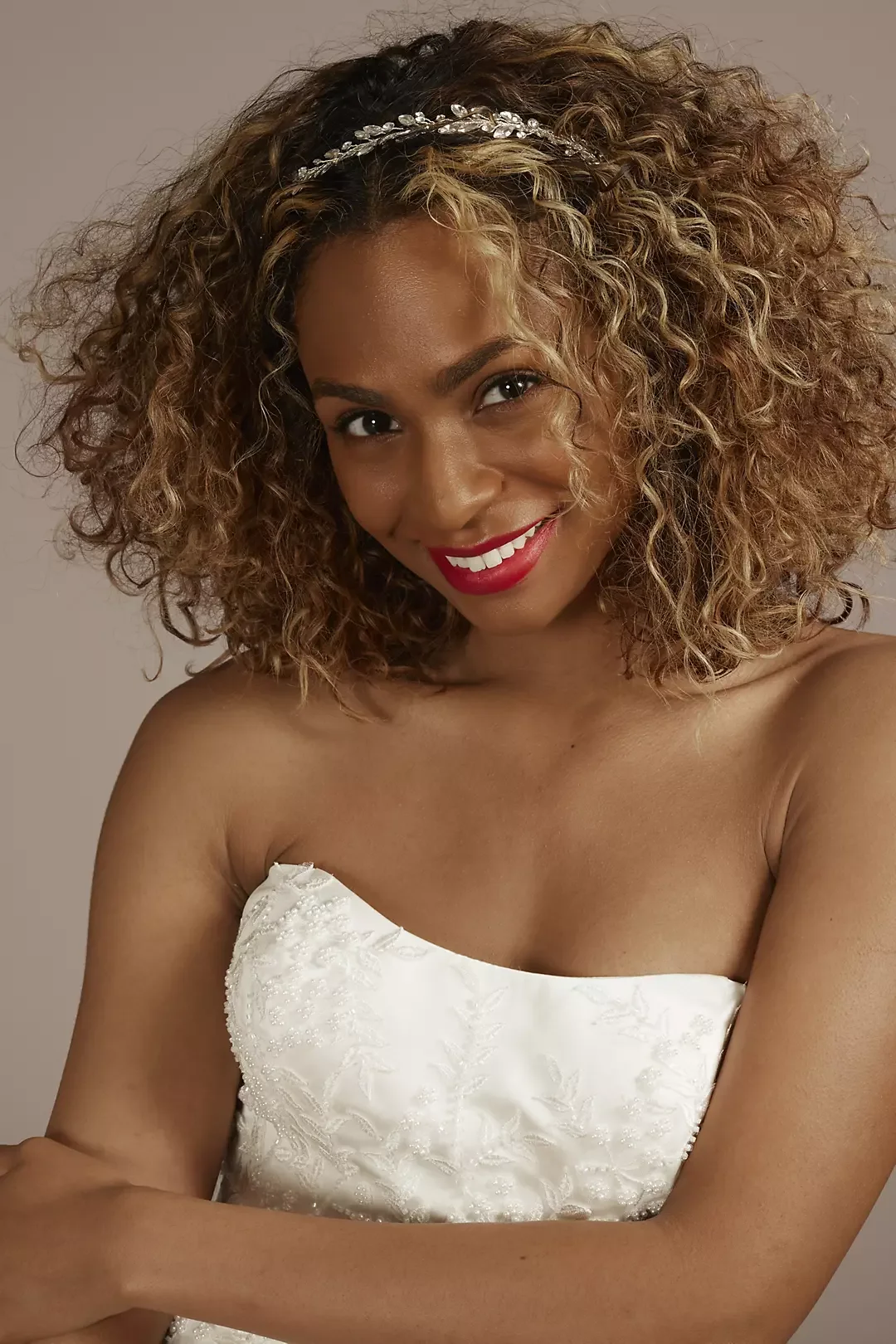 A woman with curly hair wearing a silver headband and a white strapless dress with floral embroidery, smiling with red lipstick, against a plain background.