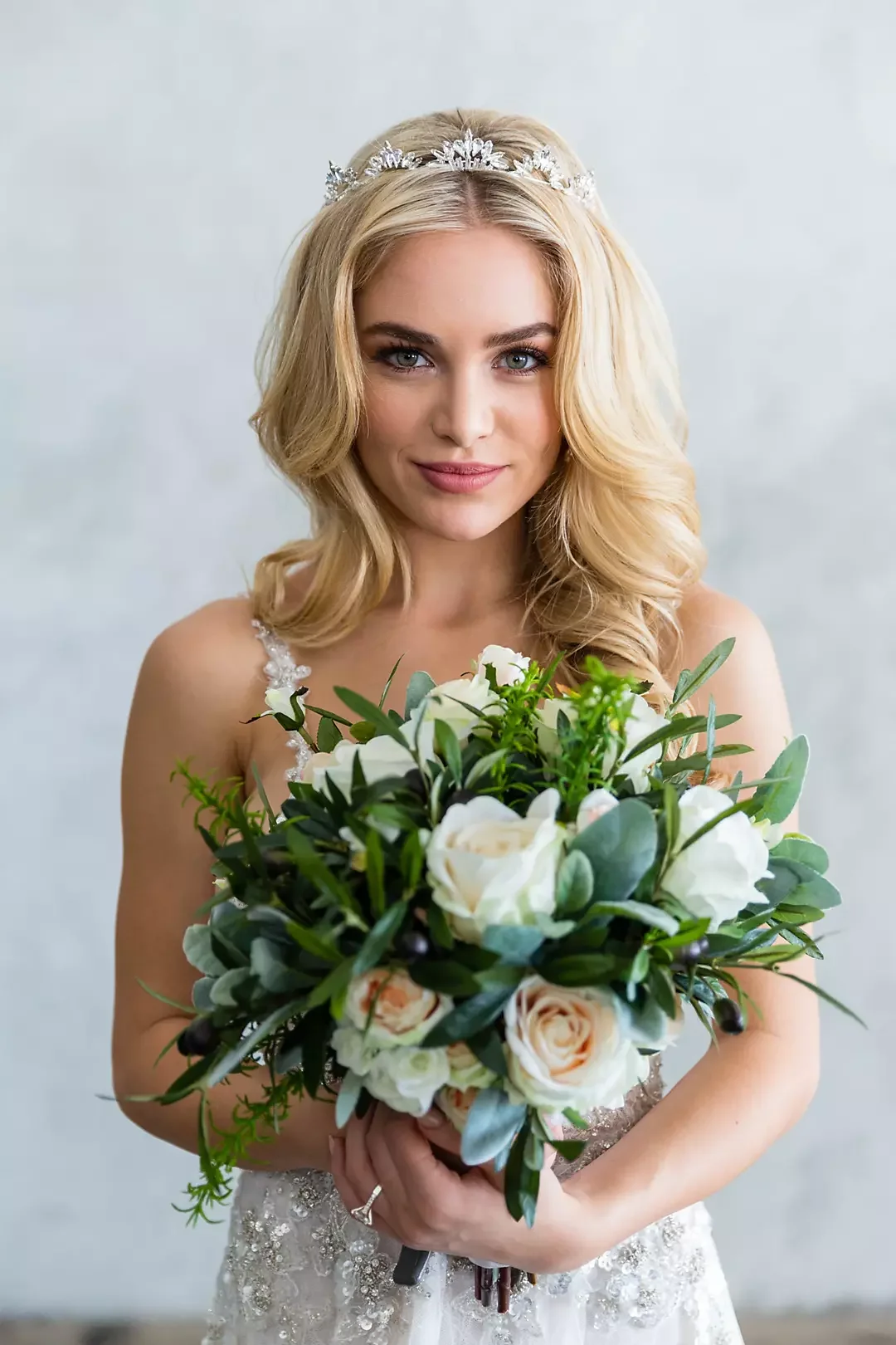 A woman with blonde hair wearing a silver tiara and a wedding dress, holding a bouquet of white and cream roses and greenery, smiling softly at the camera.