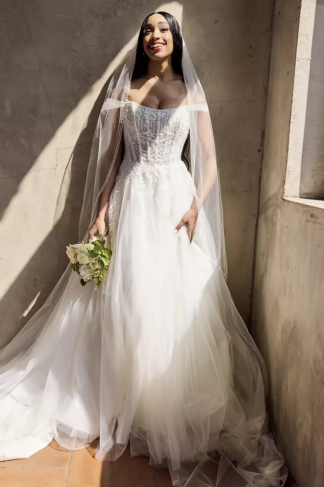 A woman in a wedding dress holding a bouquet, standing against a concrete wall, smiling.