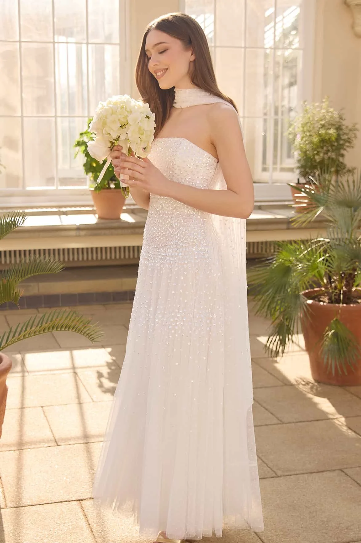 A young woman in a white, beaded, strapless dress and a matching choker holds a bouquet of white flowers. She stands indoors near large windows with natural light, surrounded by potted plants.