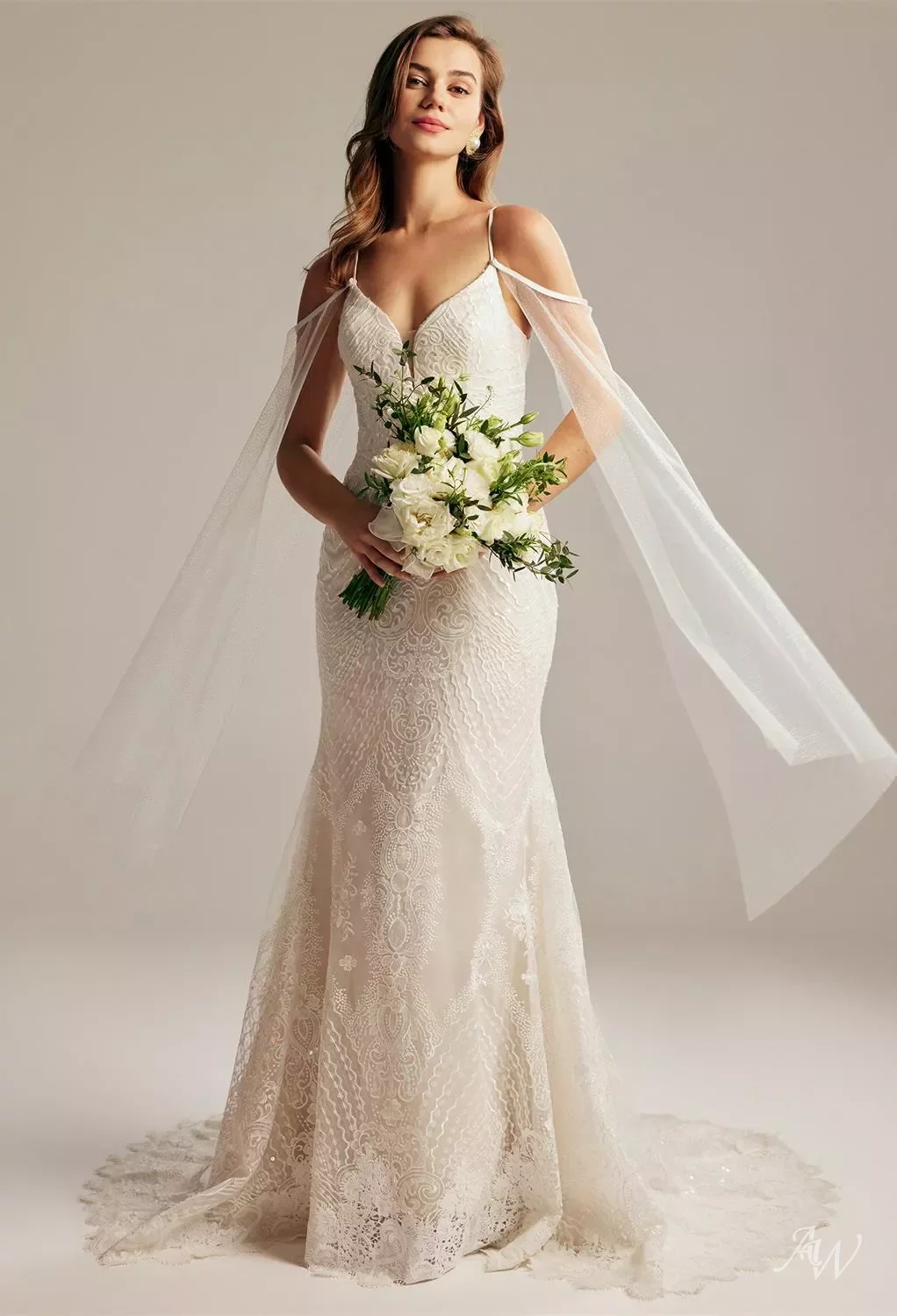 Bride in a white lace wedding gown holding a bouquet of white flowers, standing against a plain background.