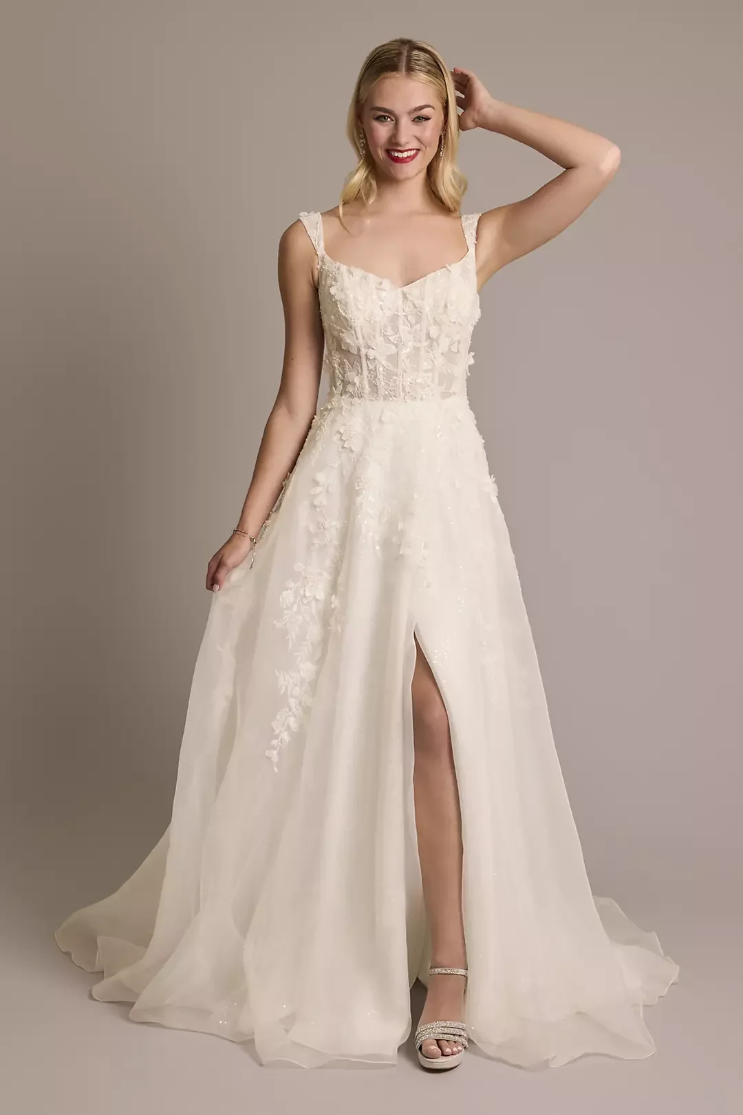 Young woman in a white wedding dress with floral embroidery, smiling, standing against a plain background.