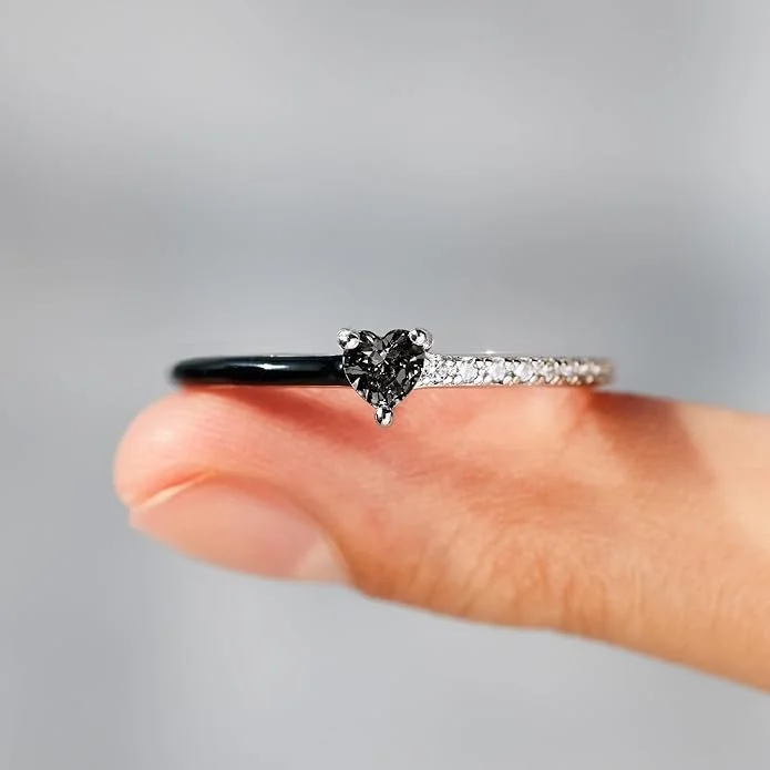 Close-up of four fingers holding two rings: a black paired ring and a silver solitaire engagement ring with a heart-shaped diamond. The background is blurred.