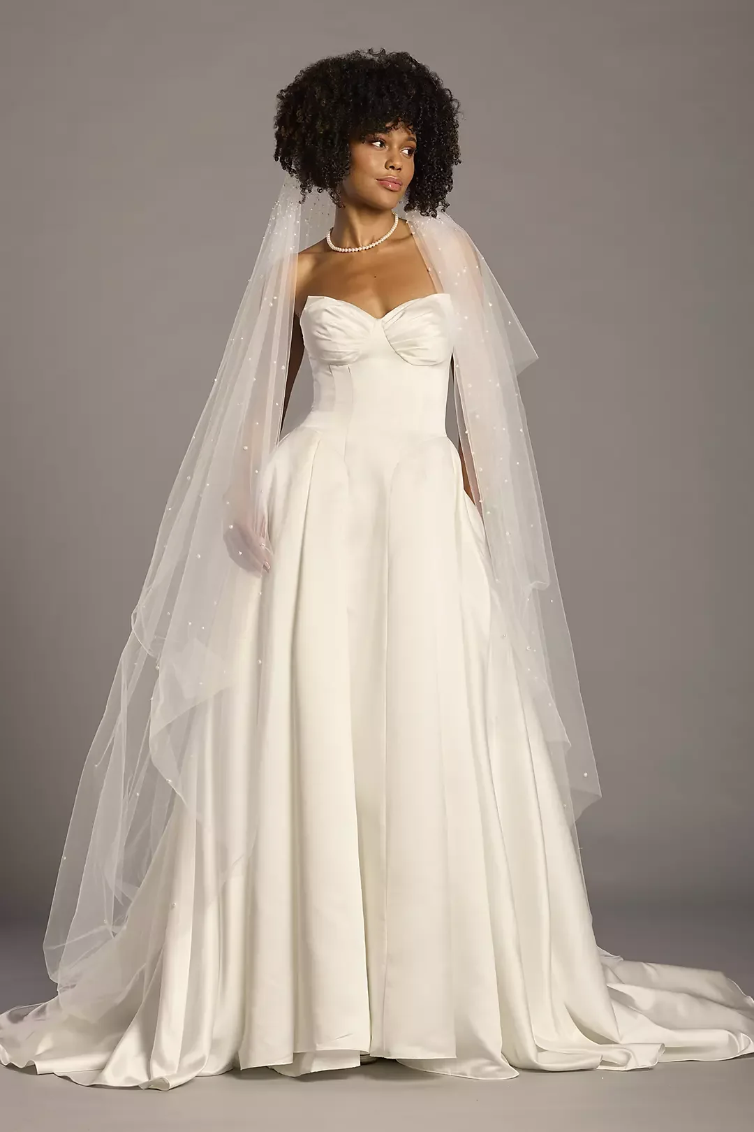 A woman in a wedding dress with a veil, pearl necklace, and short curly hairstyle stands against a neutral gray background.