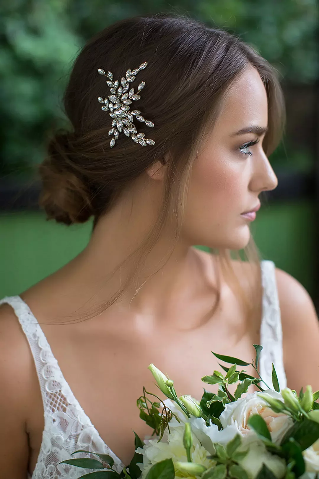 A woman with brown hair styled in an elegant updo, wearing a jeweled hair accessory, holding a bouquet of white and green flowers, wearing a lace white dress, outdoors with blurred greenery in the background.
