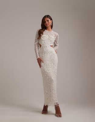 A woman wearing a white lace long-sleeve gown with cutouts at the waist, standing against a plain gray background.