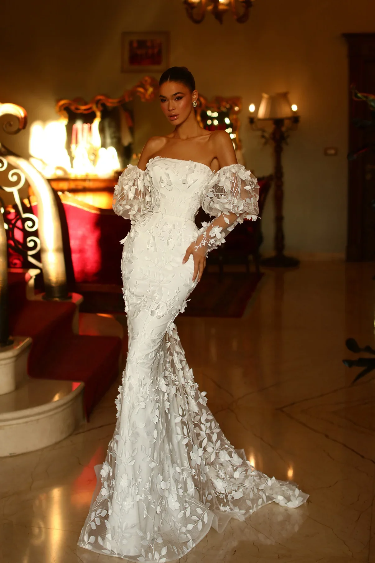 A woman in a fitted white wedding dress with floral appliqué details and off-the-shoulder puffed sleeves standing indoors in a warmly lit room.