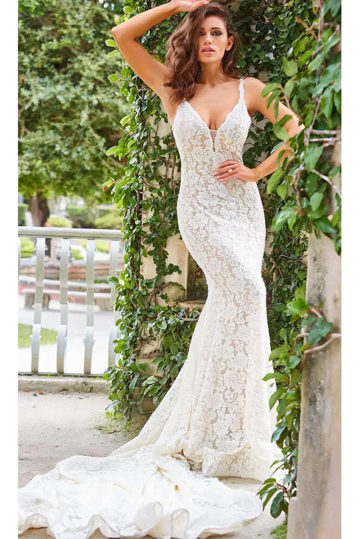 A woman in a white lace wedding dress with a V-neckline and spaghetti straps, standing outdoors against a backdrop of green vines and trees, with her hand resting on her head and her other hand on her hip.