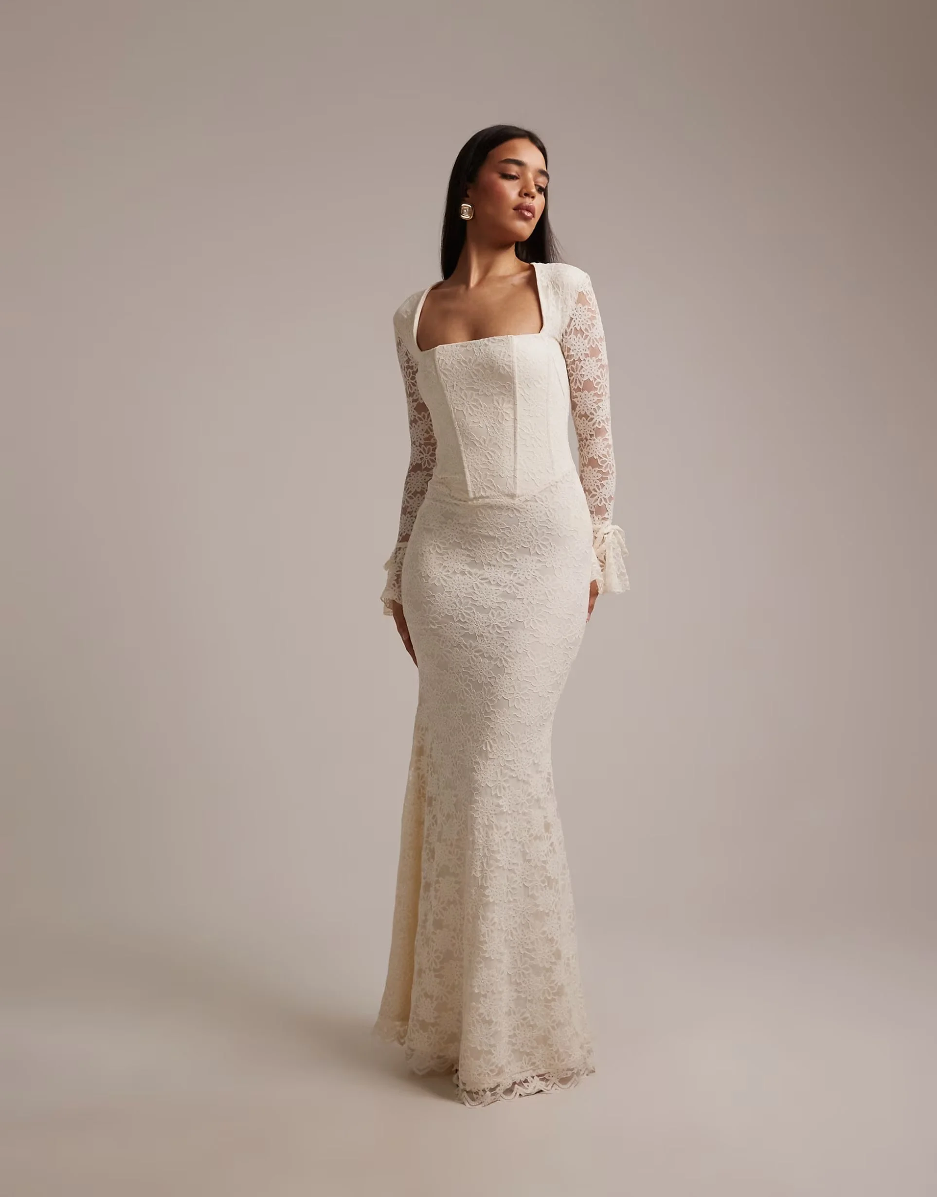 A woman wearing an elegant, long, cream-colored lace dress with long sheer lace sleeves and a square neckline, standing against a plain gray background.