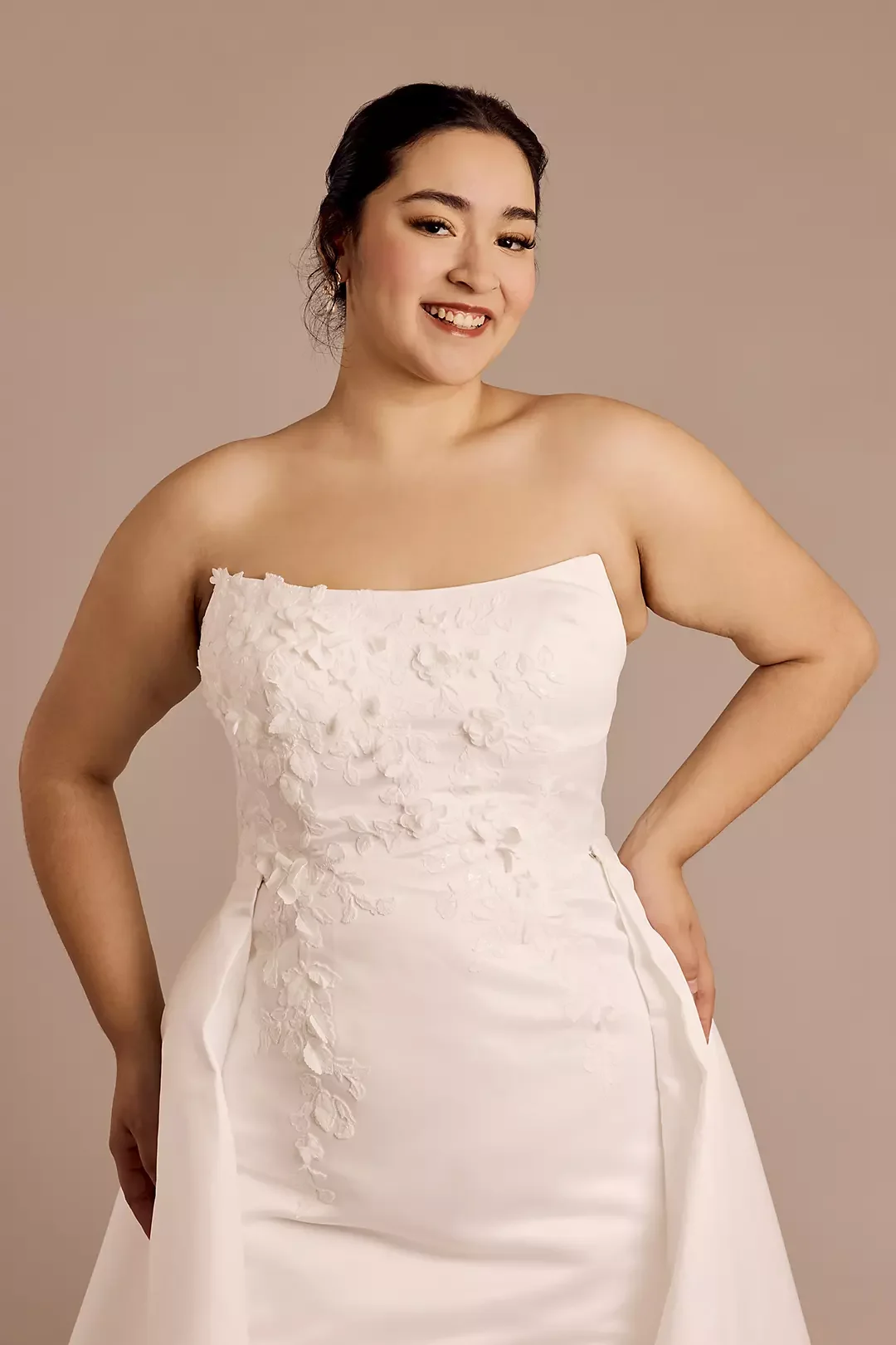 A woman smiling and wearing a strapless white wedding dress with floral embroidery.