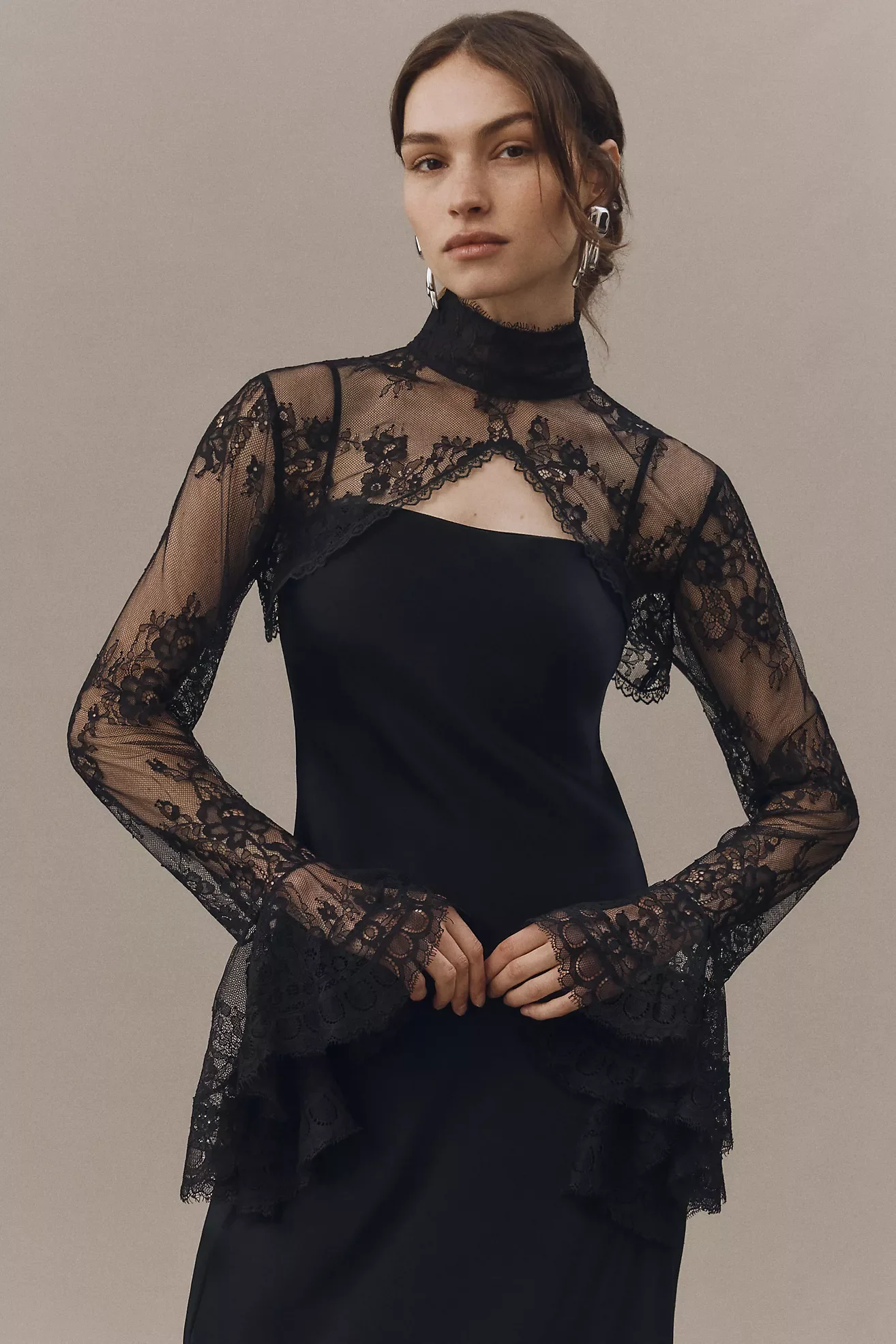 A woman wears a black dress with lace details on the sleeves and chest, standing against a plain background.