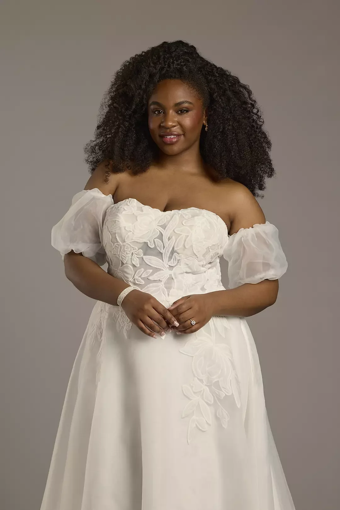 A woman with dark curly hair wearing a white off-the-shoulder wedding dress with floral embroidery, standing against a plain background.