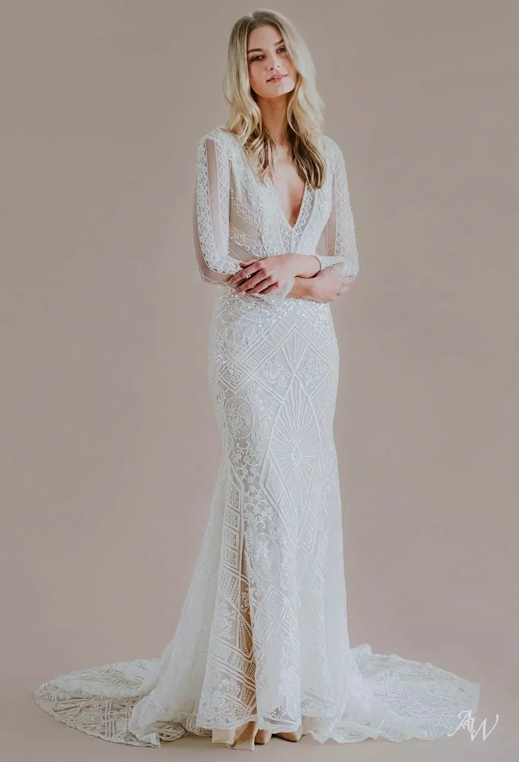 A woman in a white, intricately detailed lace wedding gown with long sleeves and a deep V neckline, standing against a plain beige background.