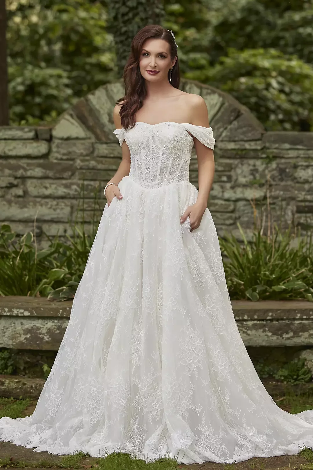 A woman in an off-the-shoulder white lace wedding dress standing outdoors in front of a stone wall and greenery.
