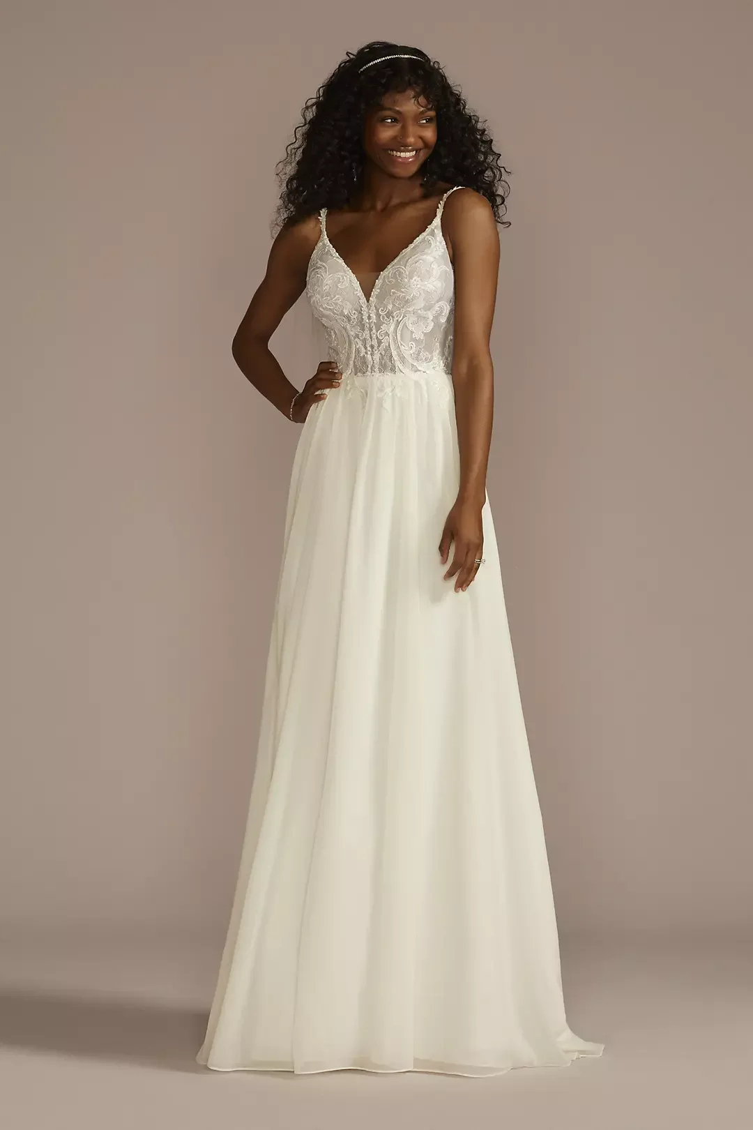 A woman in a white wedding dress with lace bodice and spaghetti straps, standing against a neutral background, smiling with one hand on her hip.