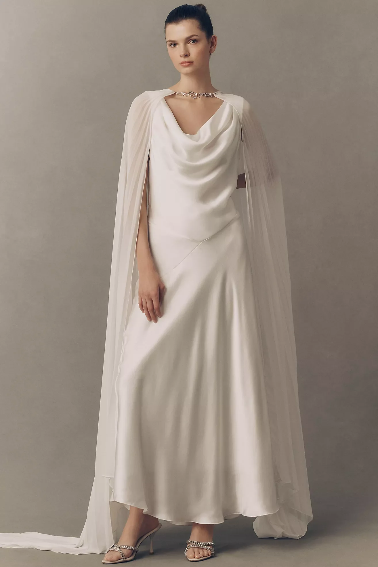 A woman wearing an elegant white satin evening gown with a draped neckline and a cape, standing against a neutral grey background. She has dark hair pulled back, and is accessorized with a silver necklace and high-heeled sandals with rhinestone details.