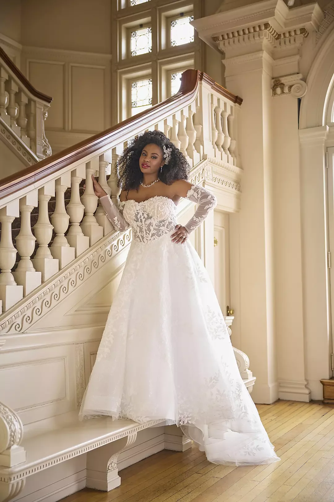 A woman in a white wedding dress with lace detailing, long lace sleeves, and a full skirt, standing on a grand staircase with ornate white woodwork and large windows behind her.
