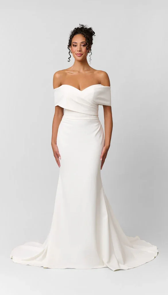 A woman wearing an off-the-shoulder white wedding gown with a fitted bodice and a long, flowing skirt, standing against a plain light gray background.