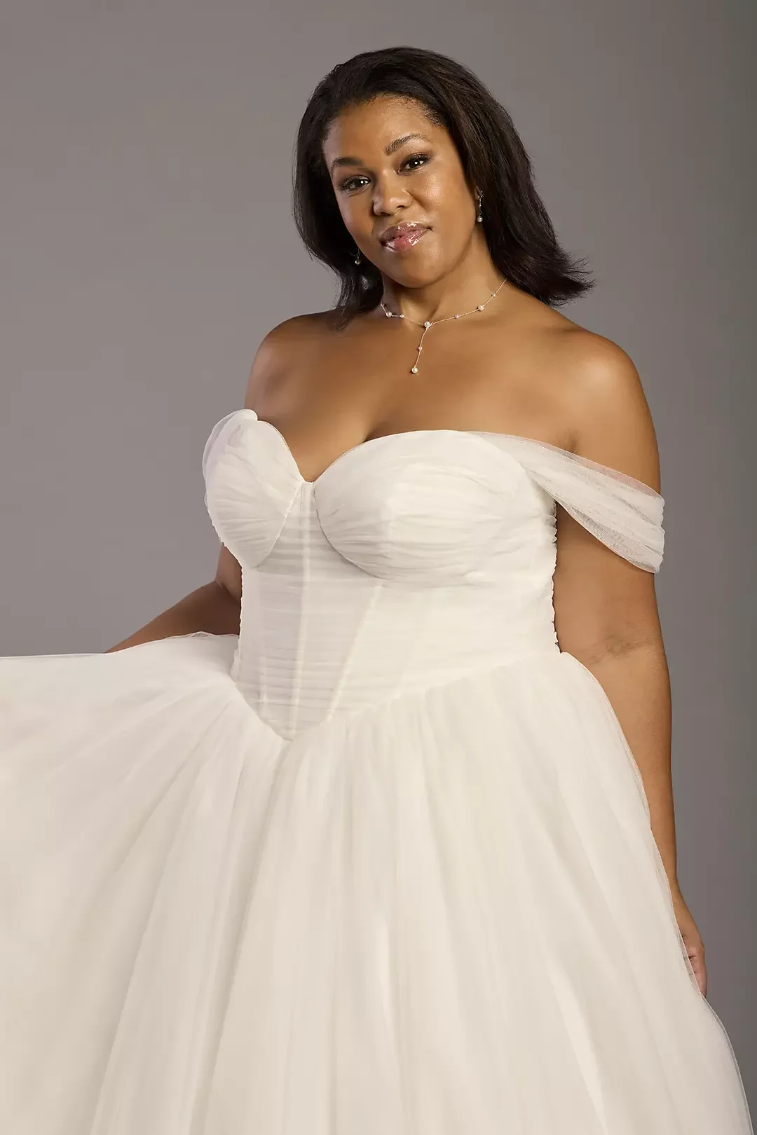 A woman in an off-shoulder white gown with a pleated bodice and a full skirt posing against a plain gray background.