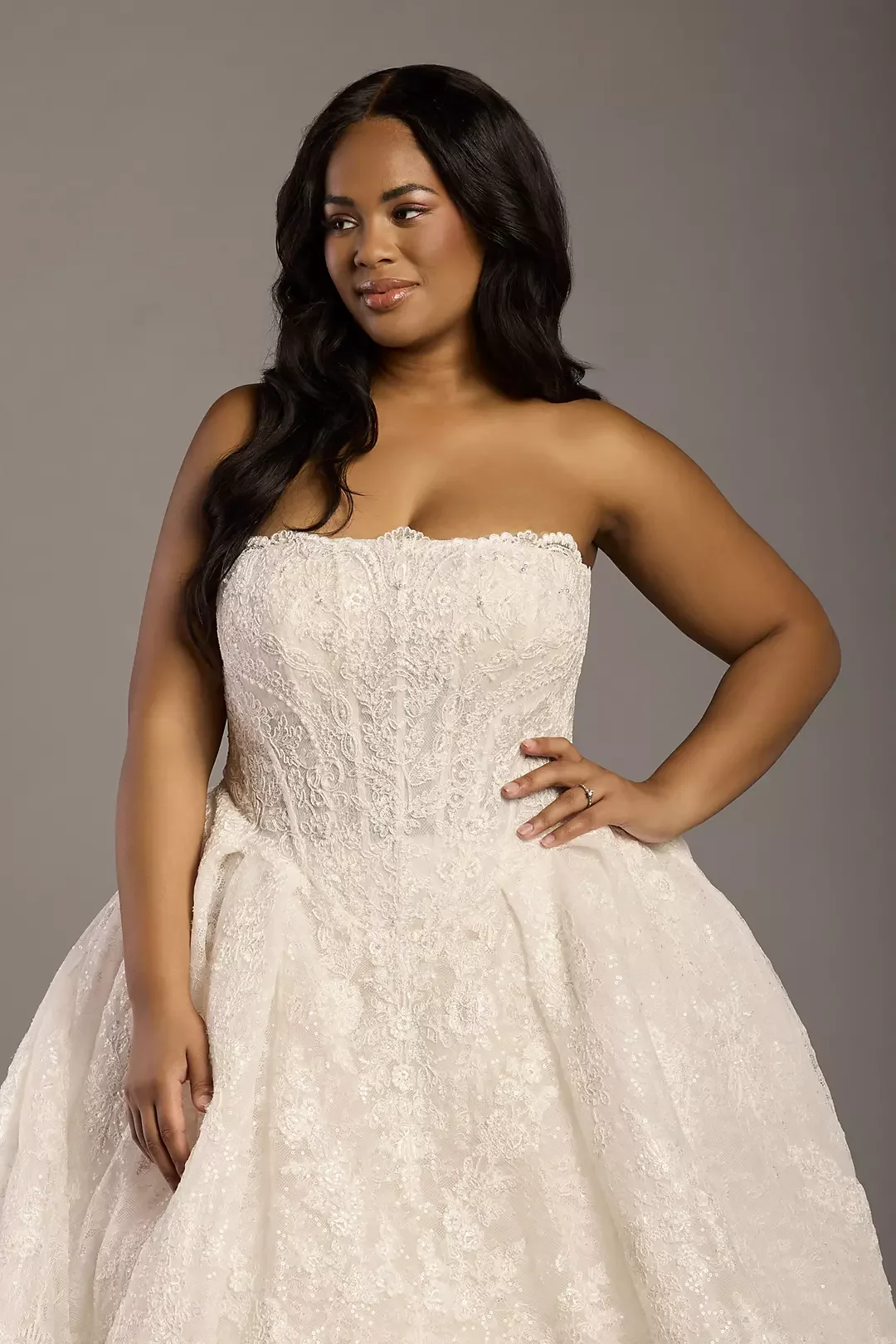 A woman in a white wedding dress with lace details, posing with one hand on her hip against a gray background.