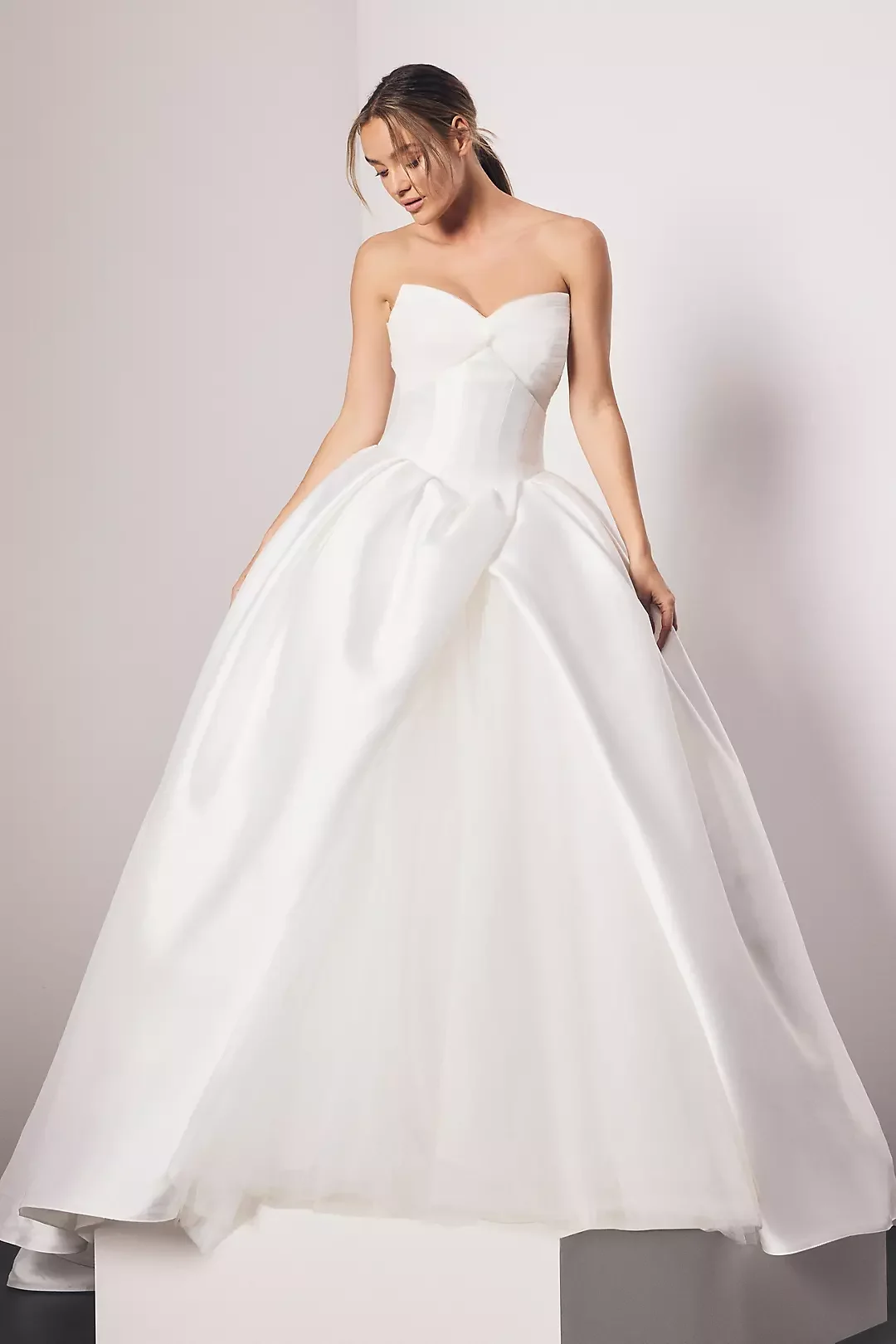 A woman wearing a strapless white wedding gown with a full skirt, standing against a plain background.