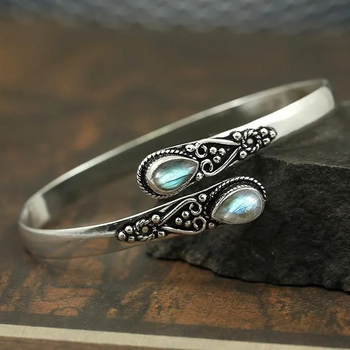 Silver bracelet with two teardrop-shaped moonstone cabochons and intricate scrollwork, resting on a dark stone on a wooden surface.